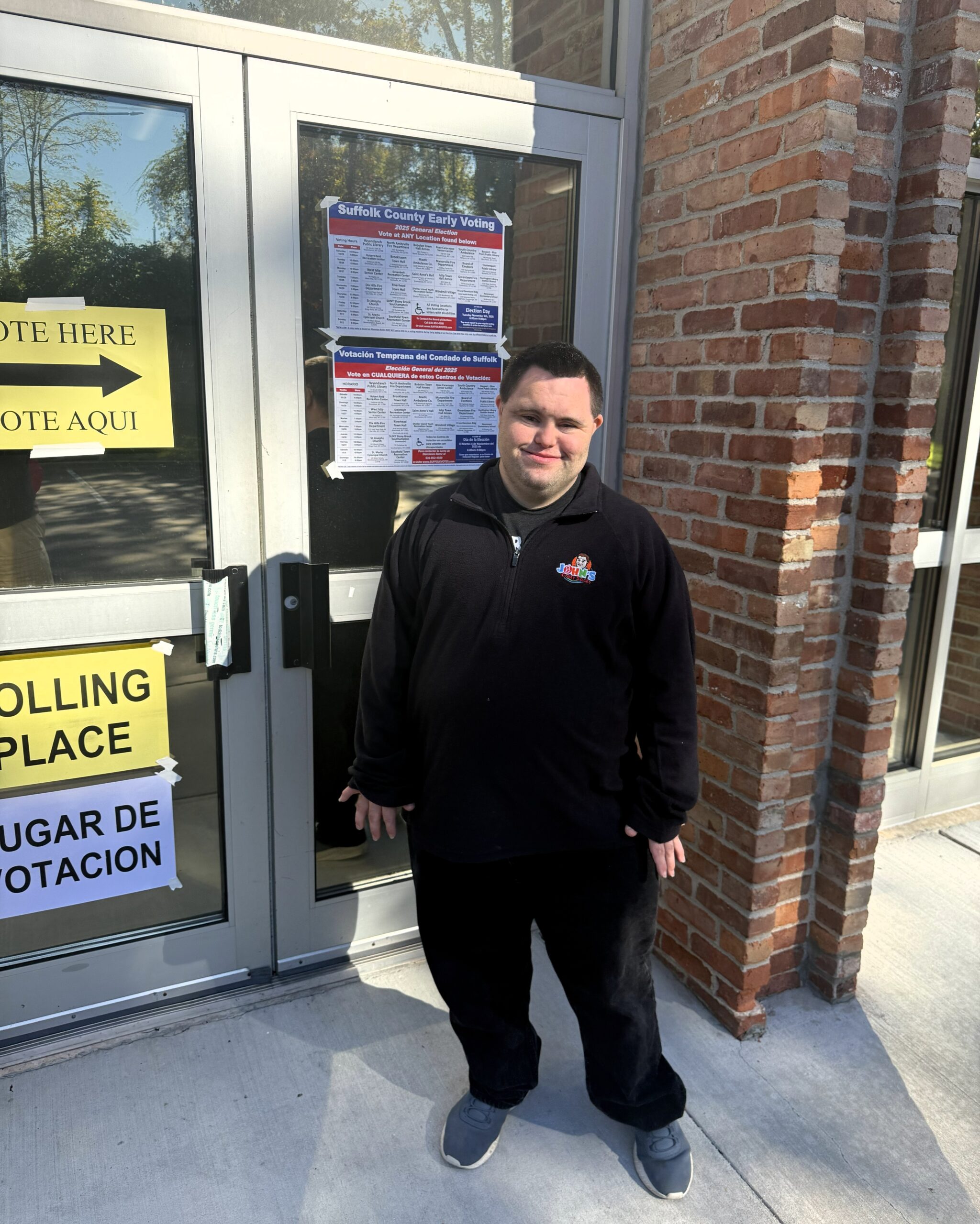 John wears a block top and black pants. He stands a the doors to the polling place, There are signs on the door marking it as a poling location.